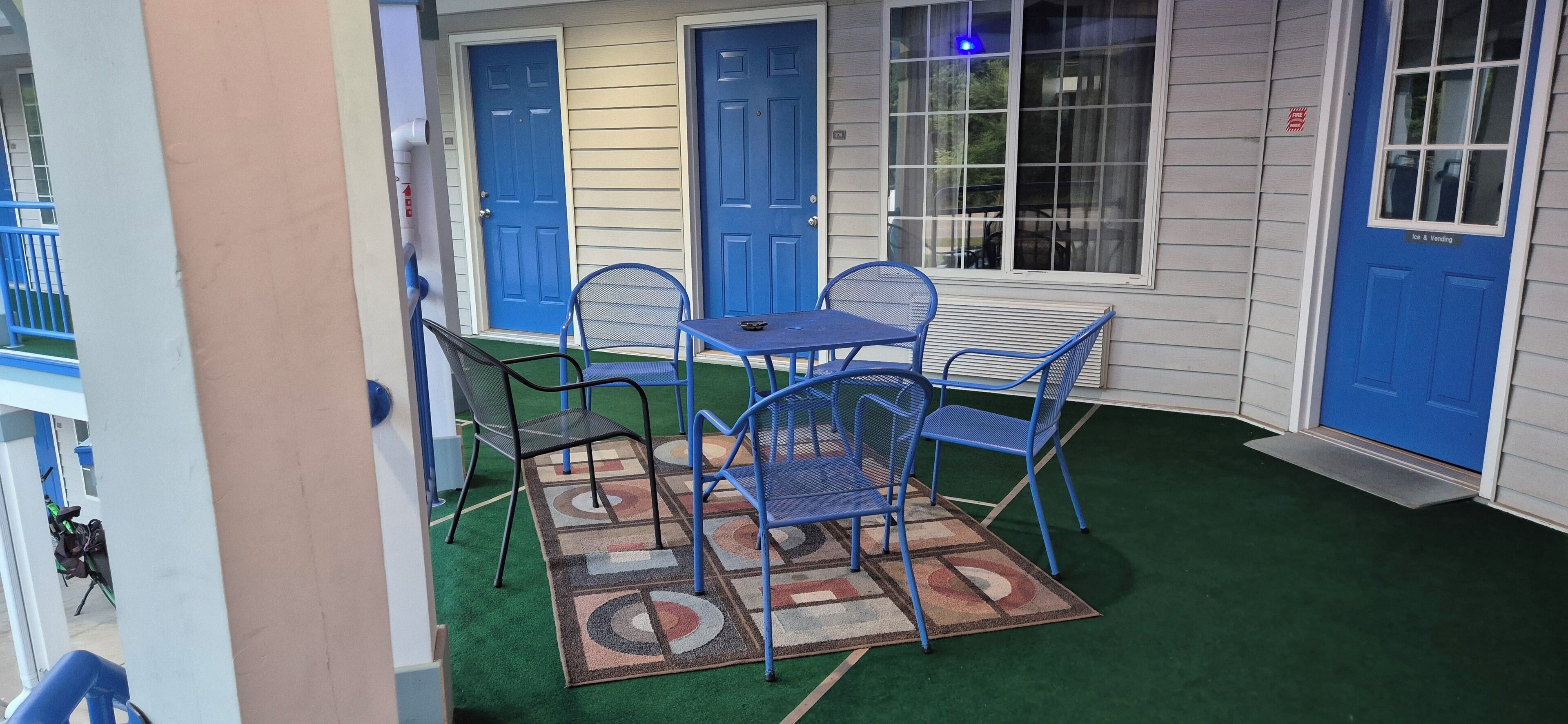A small outdoor seating area with blue metal chairs and a table, a blue door, and a window with a view of the outside.