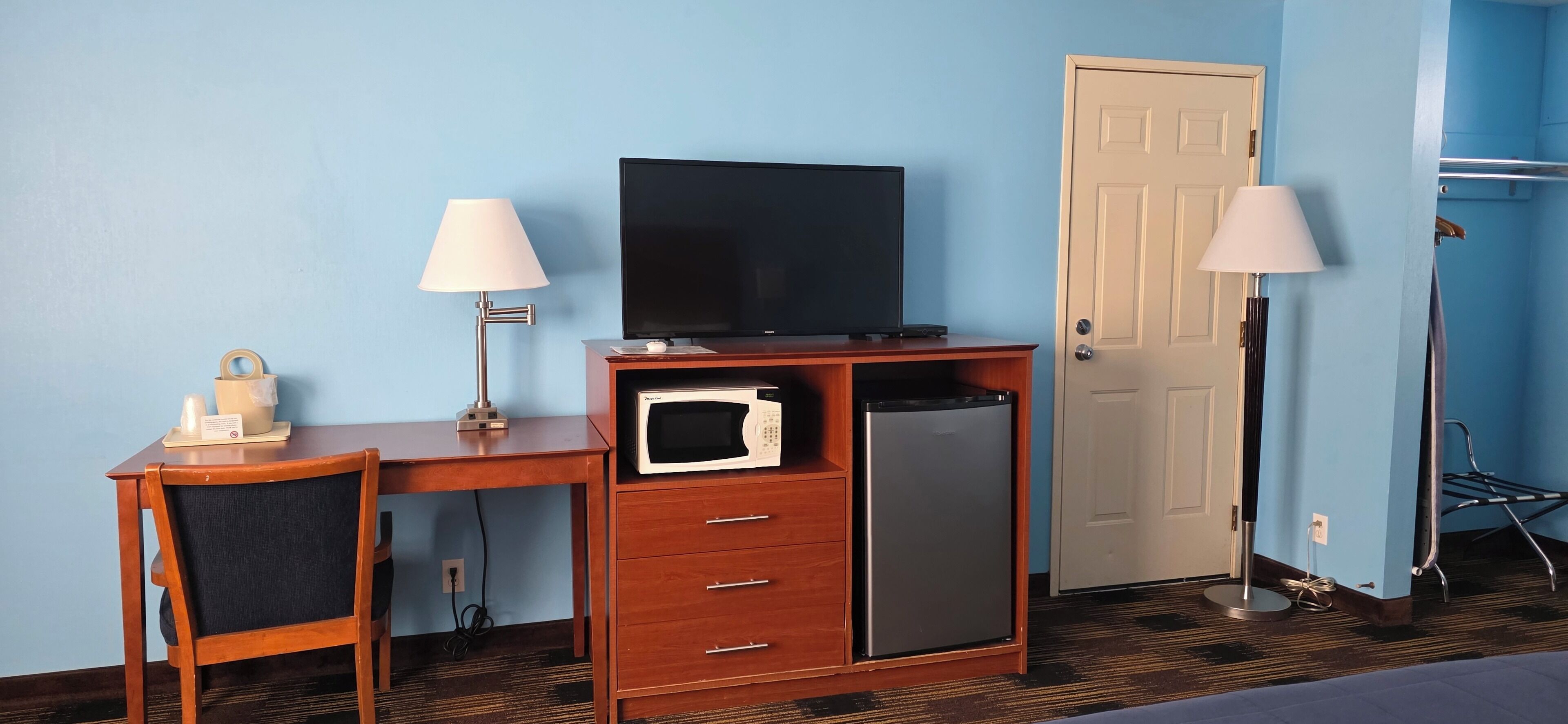 A hotel room with a wooden desk, a television, a microwave, a mini-fridge, two lamps, a door, and a coat rack.