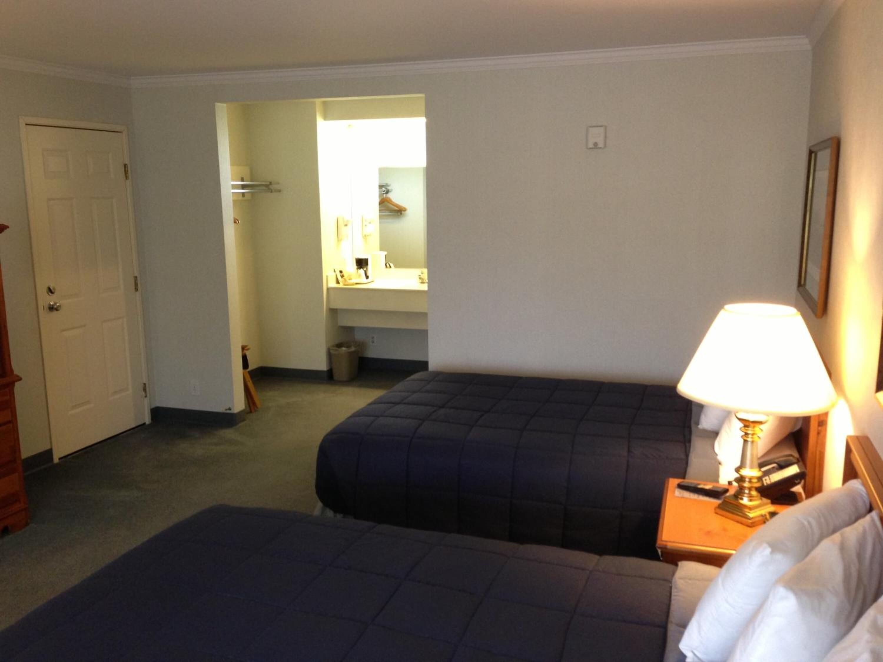 A hotel room with two beds, a bedside table with a lamp, a bathroom visible through an open door, and a coat rack.