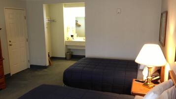 A hotel room with two beds, a bedside table with a lamp, a bathroom visible through an open door, and a coat rack.