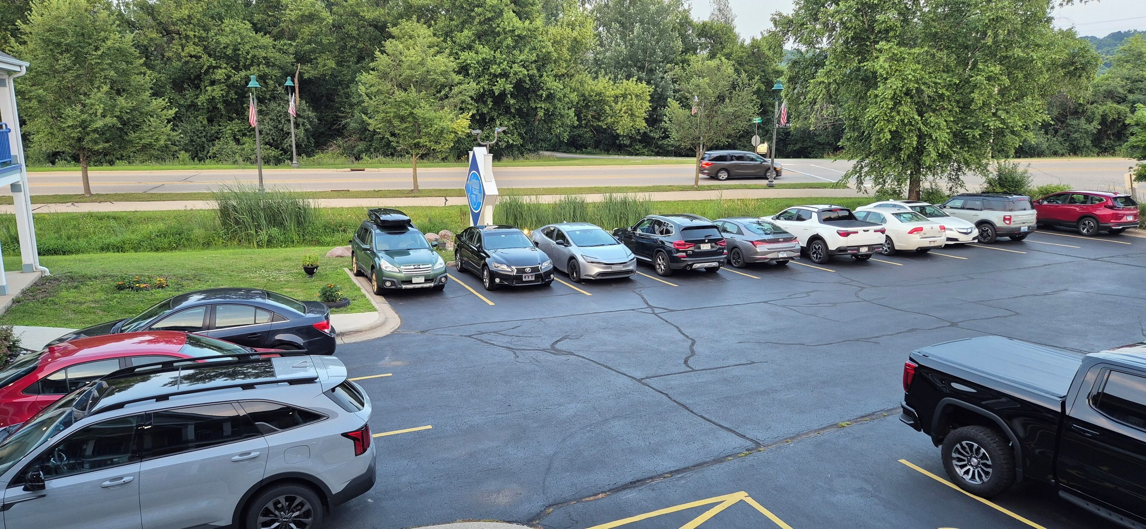 A parking lot with multiple cars parked in designated spaces, surrounded by greenery and a road in the background.