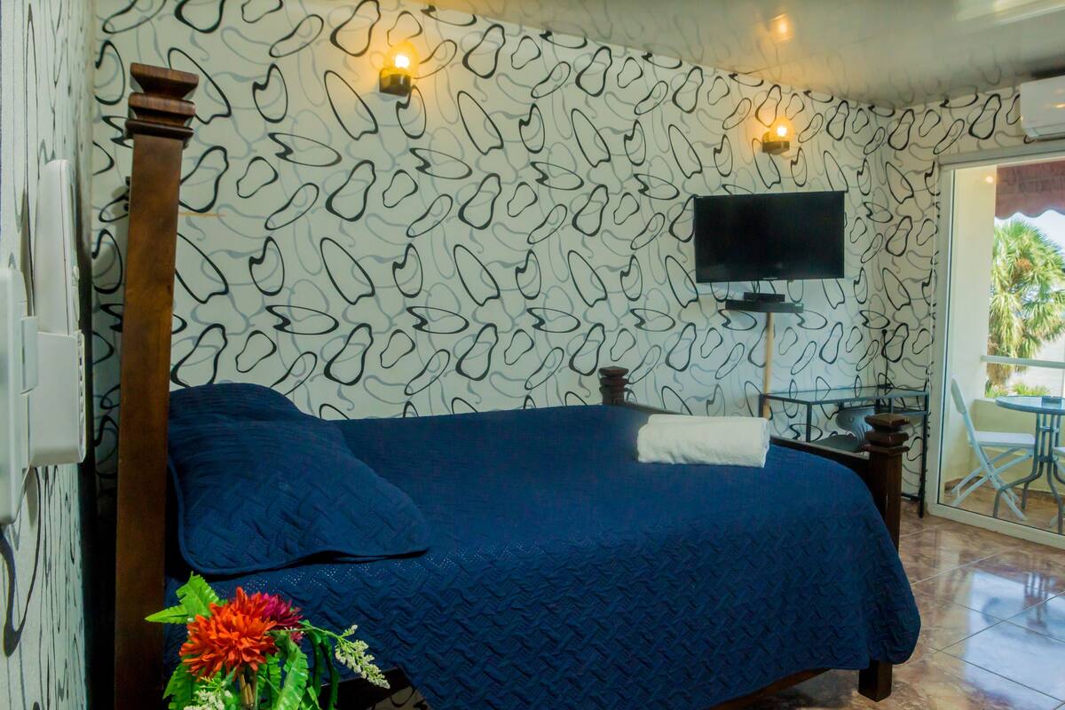 A bedroom with a bed, a TV on the wall, a small table with a chair, and a balcony with a table and chairs.
