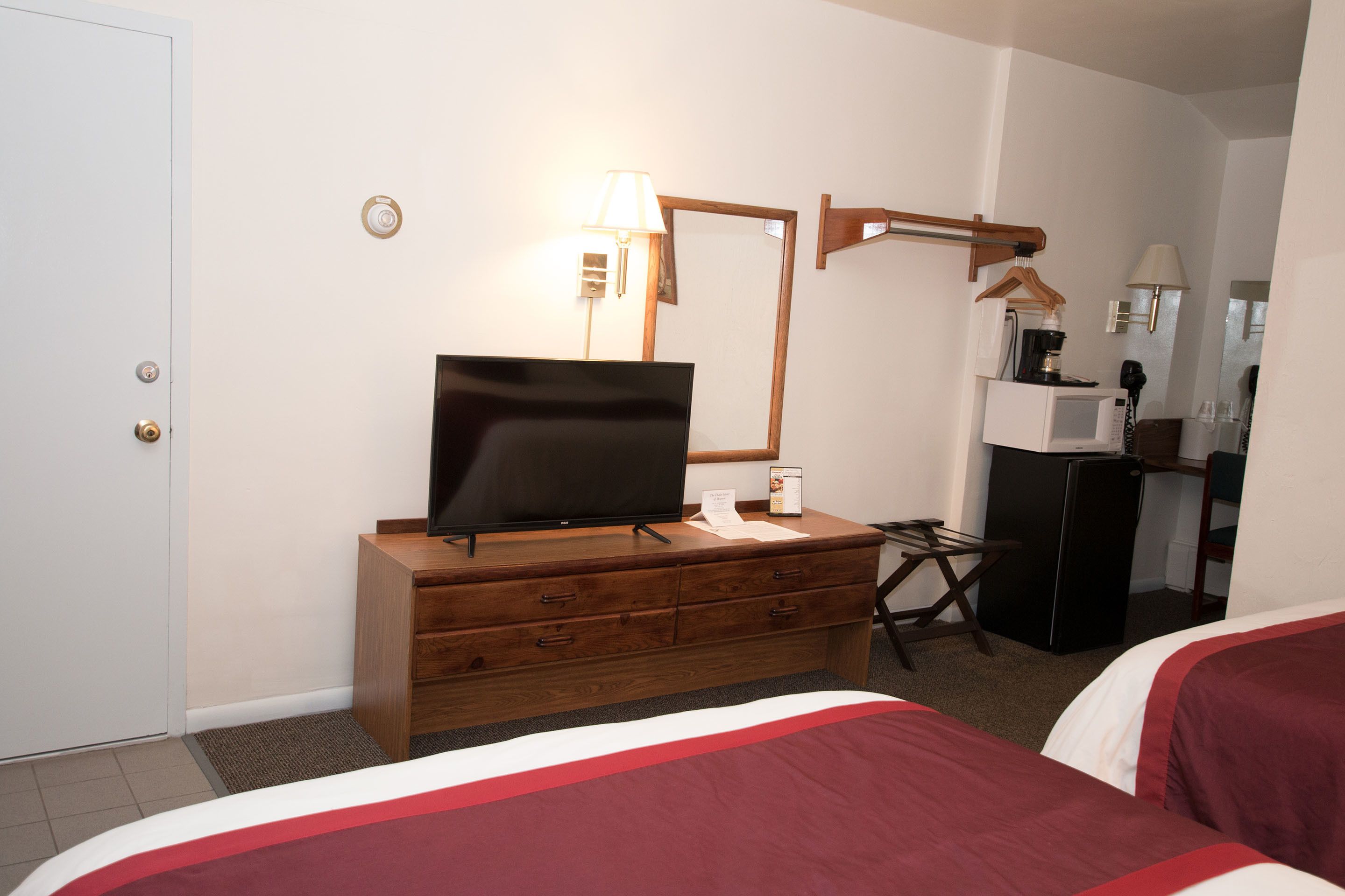A hotel room with a TV on a wooden dresser, a mirror, a desk with a lamp, a refrigerator, and two beds with red and white bedding.