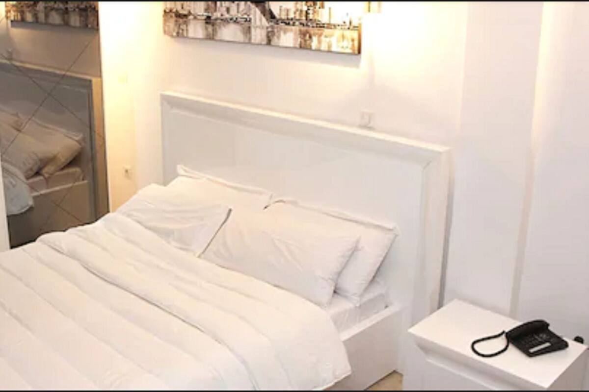 Un lit bien fait, avec une literie blanche, une table de chevet sur laquelle se trouve un téléphone noir, et une salle de bain où l’on aperçoit un porte-serviettes.