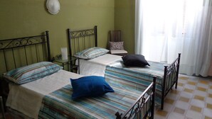 Ein Zimmer mit zwei Betten, jeweils mit einem gemusterten Überwurf und Kissen, einem Sessel mit einem gemusterten Sitzkissen und einem Fenster mit durchsichtigen Vorhängen.