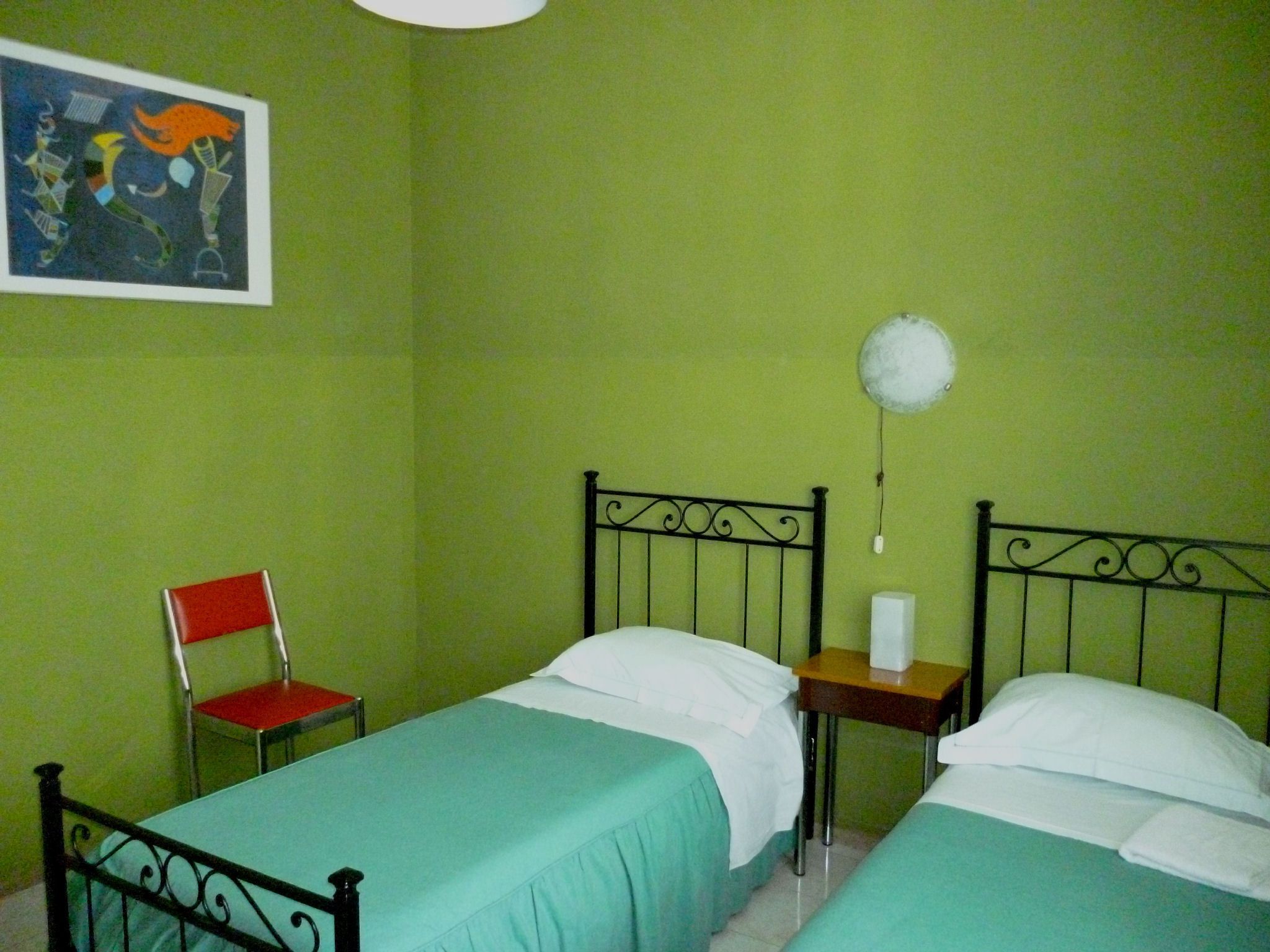 A room with two single beds, a chair, a small table, and a wall-mounted lamp.