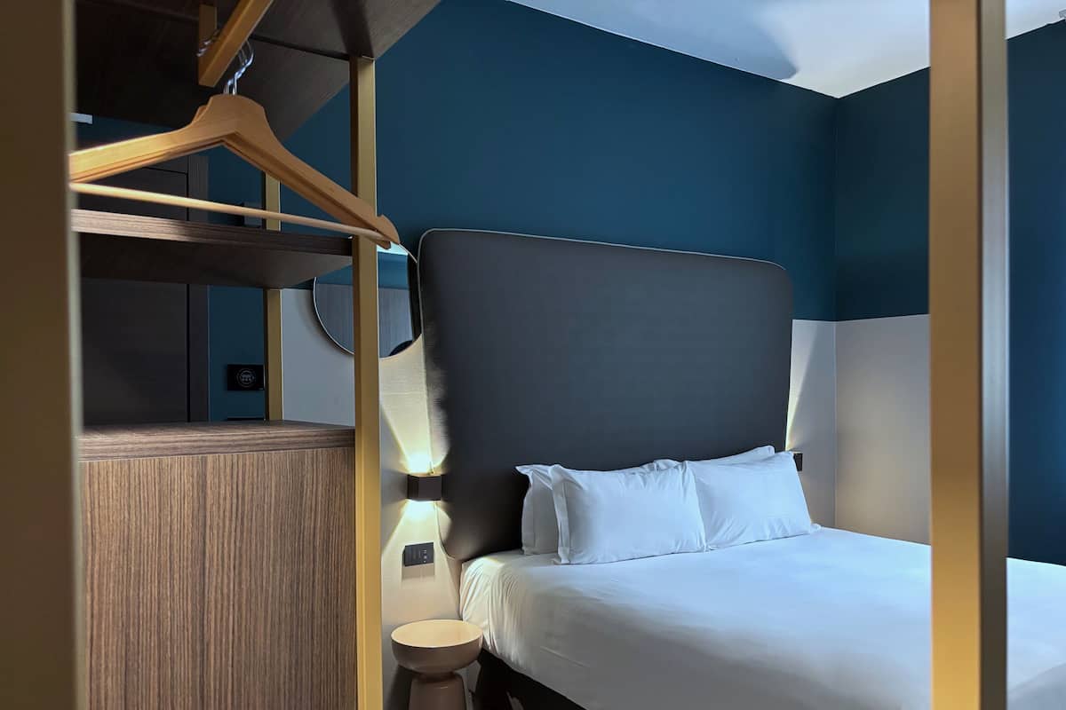 A modern hotel room with a large bed, bedside table, and a wooden wardrobe.