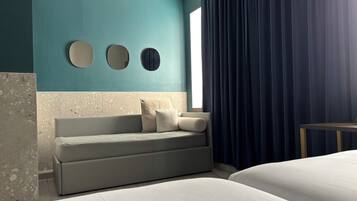 Ein modernes Hotelzimmer mit einer türkisfarbenen Akzentwand, einer Couch mit zwei Kissen, einem Bett und einem großen Fenster mit dunklen Vorhängen.