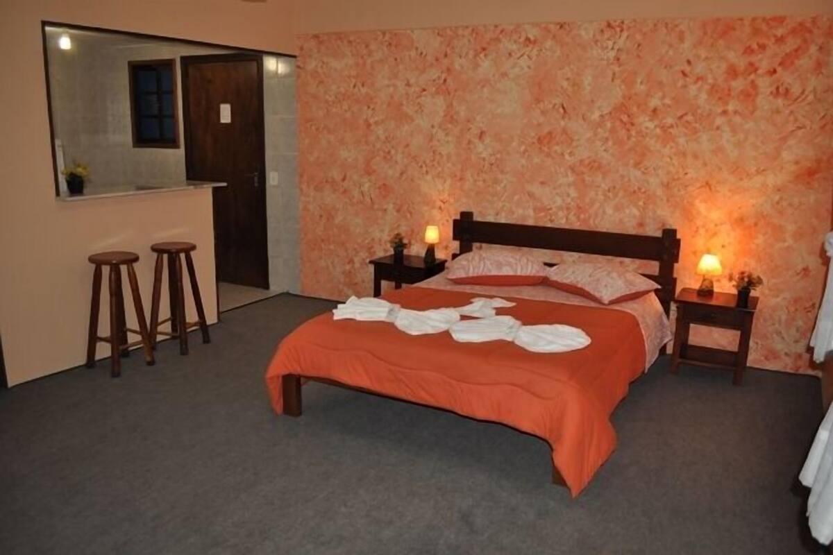 Una habitación de hotel con cama, dos mesitas de noche, un espejo y una pequeña zona de bar con taburetes.