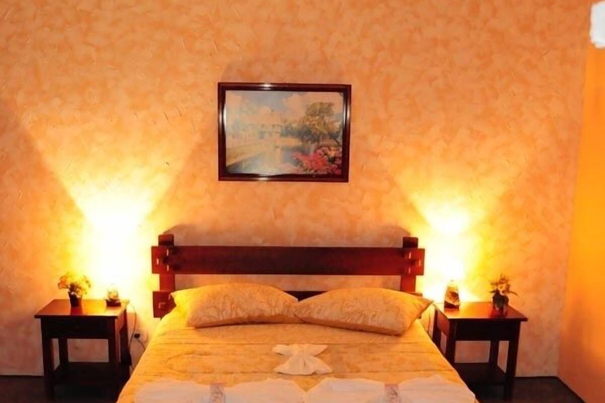 Una cama bien hecha con colcha y almohadas estampadas, con dos mesitas de noche y lámparas a cada lado, en una habitación con pared texturizada y un cuadro enmarcado sobre la cama.