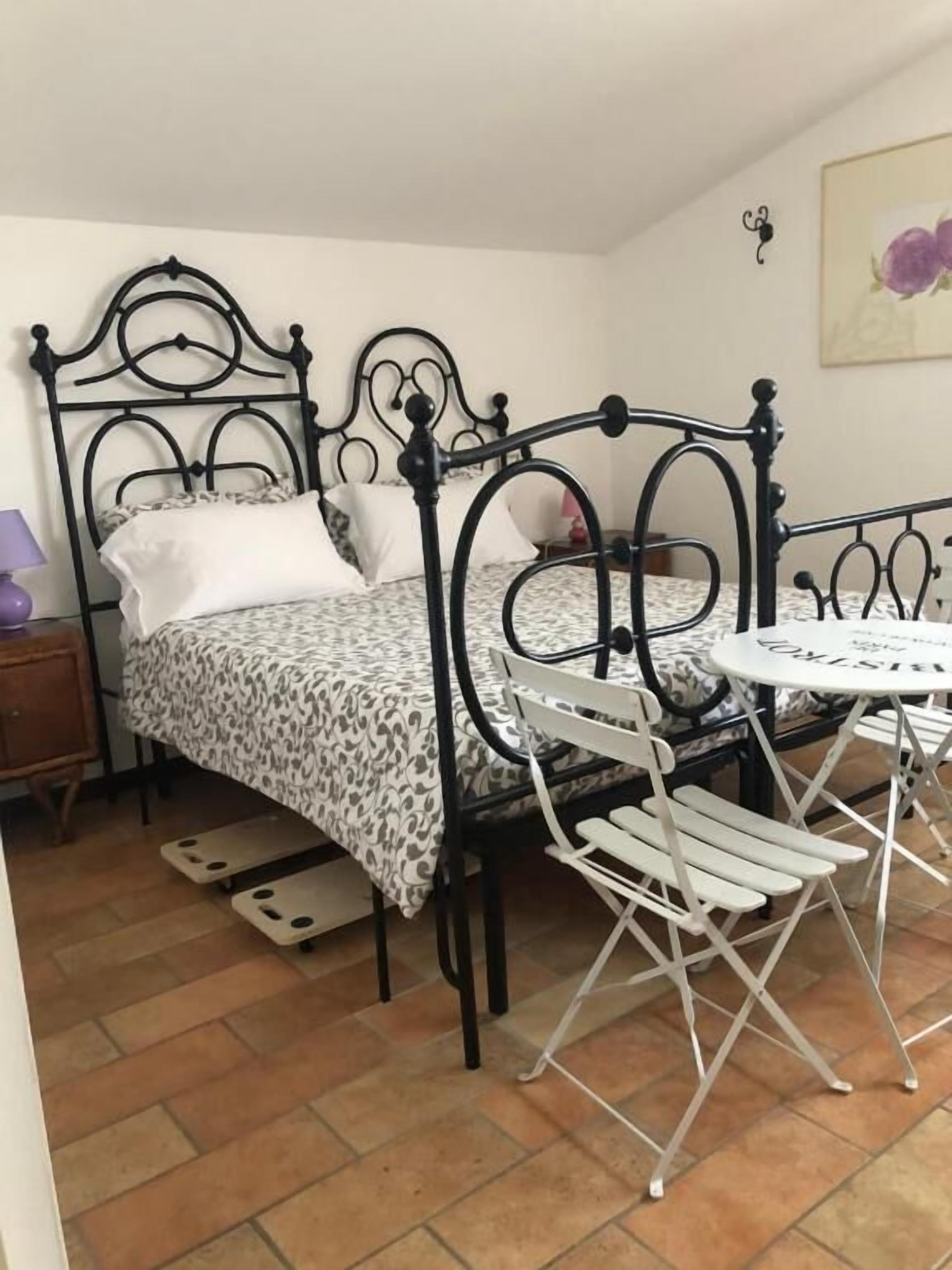 A bed with a metal frame, a small round table, and a chair in a room with tiled flooring.