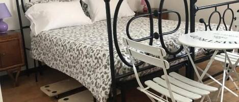 A bed with a metal frame, a small round table, and a chair in a room with tiled flooring.