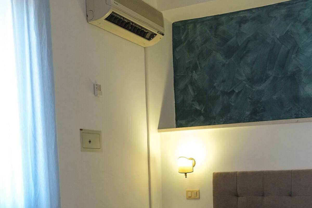 A hotel room with a bed, a wall-mounted air conditioner, a night light, and a painting.