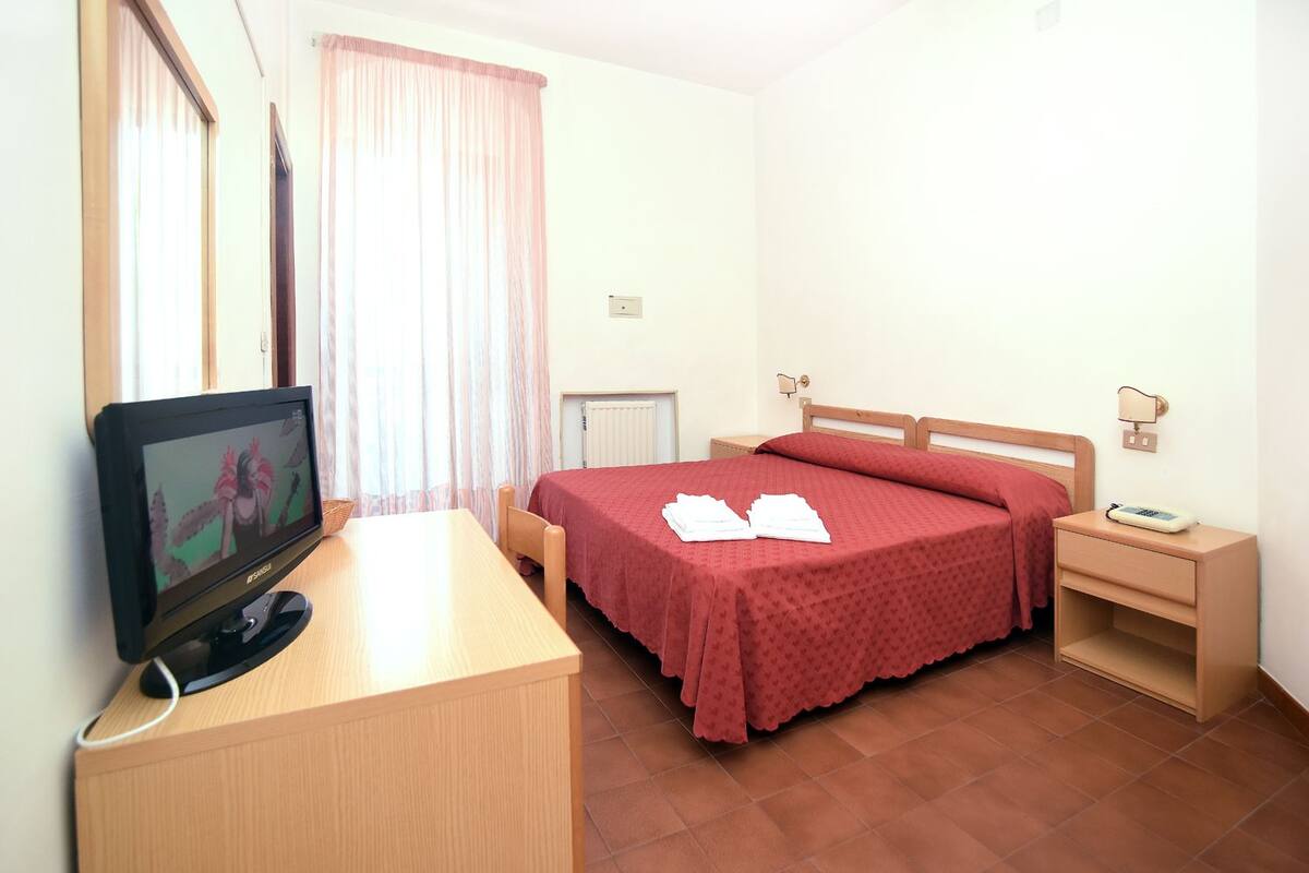 A hotel room with a bed, a bedside table, a TV, and a window with curtains.