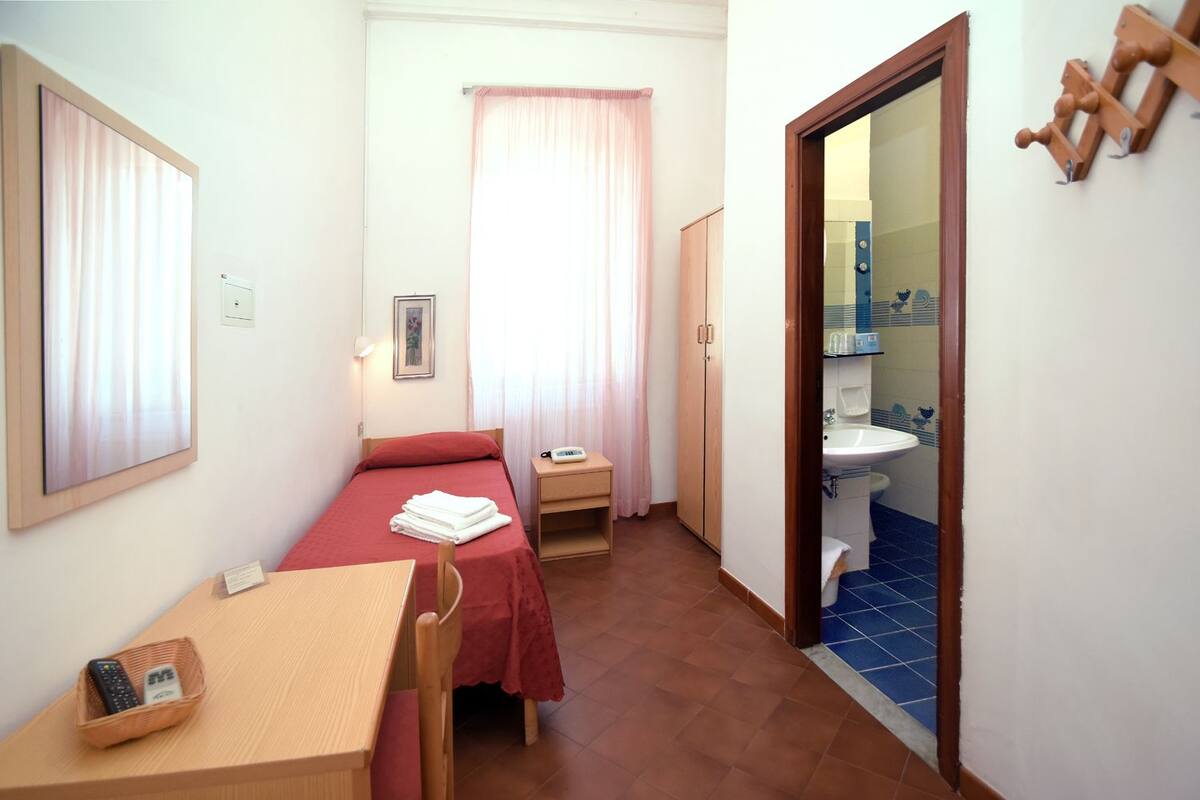 A small, compact room with a single bed, a bedside table with a lamp, a chair, a small table with a basket, and a bathroom with a sink and toilet visible through an open door.