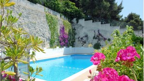 Una piscina con plataforma blanca, rodeada de plantas y flores.