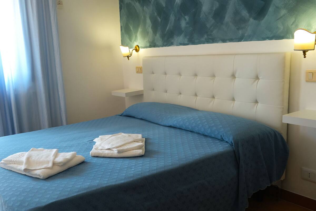 A neatly made bed with a blue bedspread and two folded white towels, a tufted white headboard, and two wall-mounted bedside lamps.