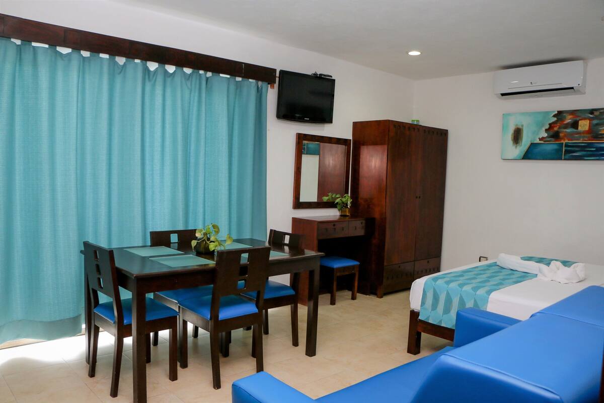 Quarto de hotel com mesa de jantar, cadeiras, cama, televisão, guarda-roupa e um quadro na parede.