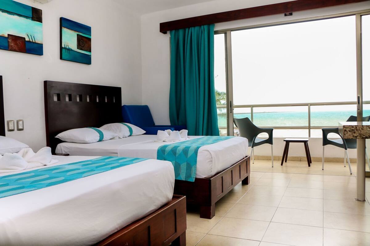 Quarto de hotel com duas camas, varanda com vista para o mar e área de refeições com cadeiras.