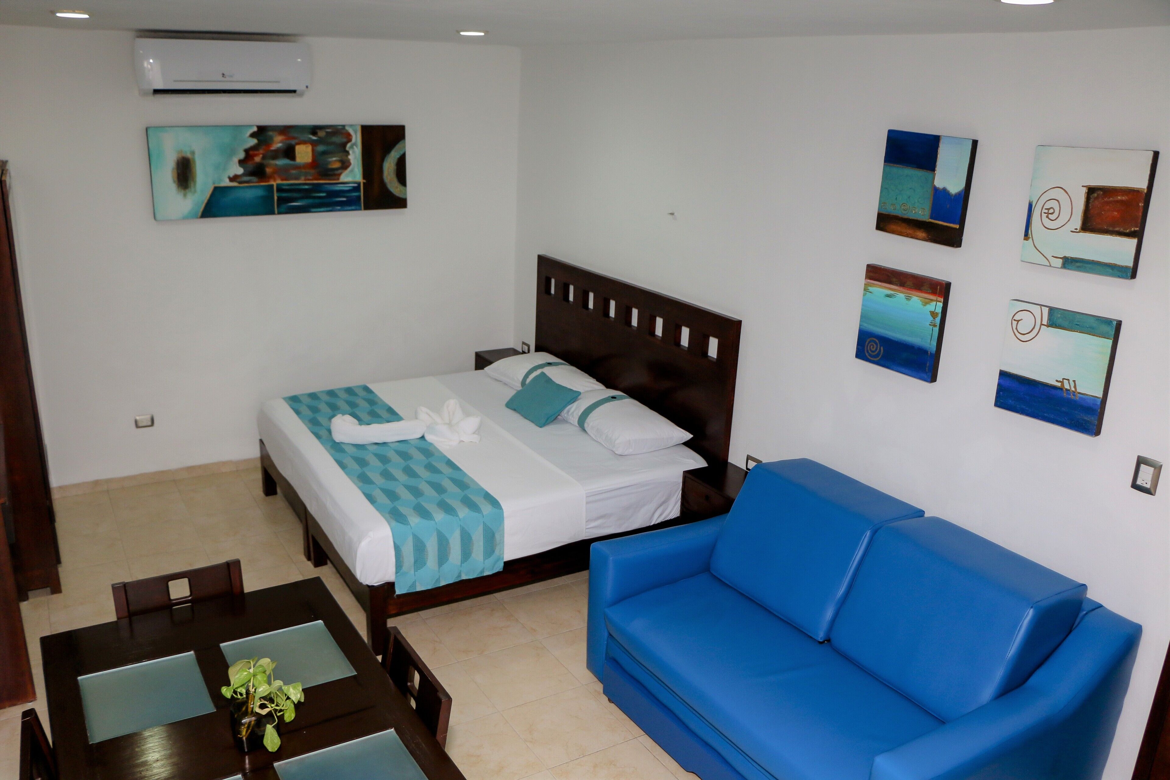 A hotel room with a bed, a blue sofa, a dining table, and artwork on the walls.