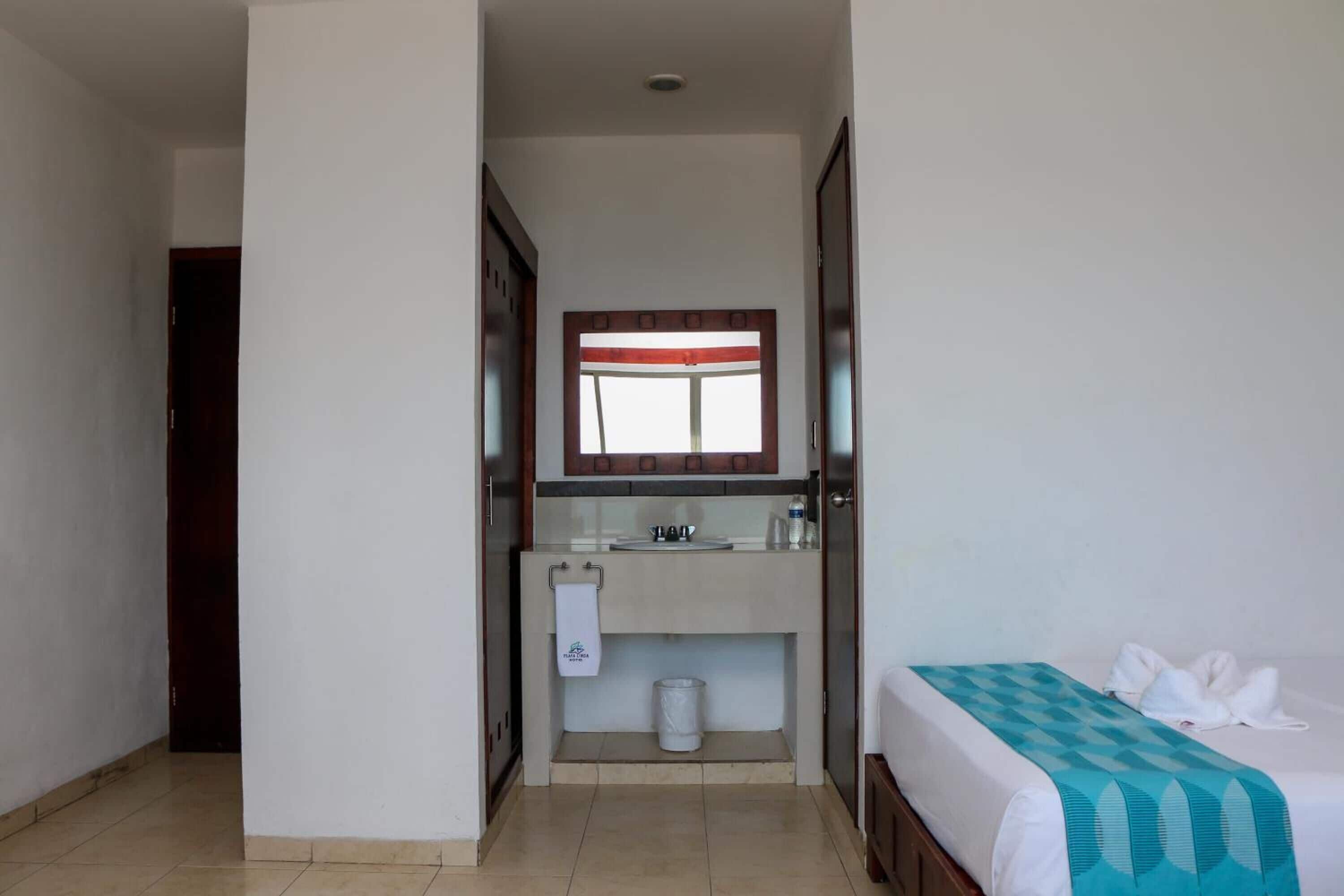 Habitación de hotel con cama, baño con lavamanos y espejo, y una puerta que da a otra habitación.