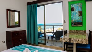 Habitación de hotel con cama, escritorio, silla, cómoda y vistas a la playa y al océano a través de una puerta corrediza de vidrio.