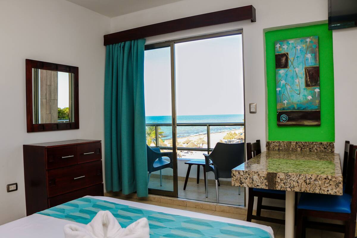 Quarto de hotel com cama, escrivaninha, cadeira, cômoda e vista para a praia e oceano através de uma porta de vidro deslizante.