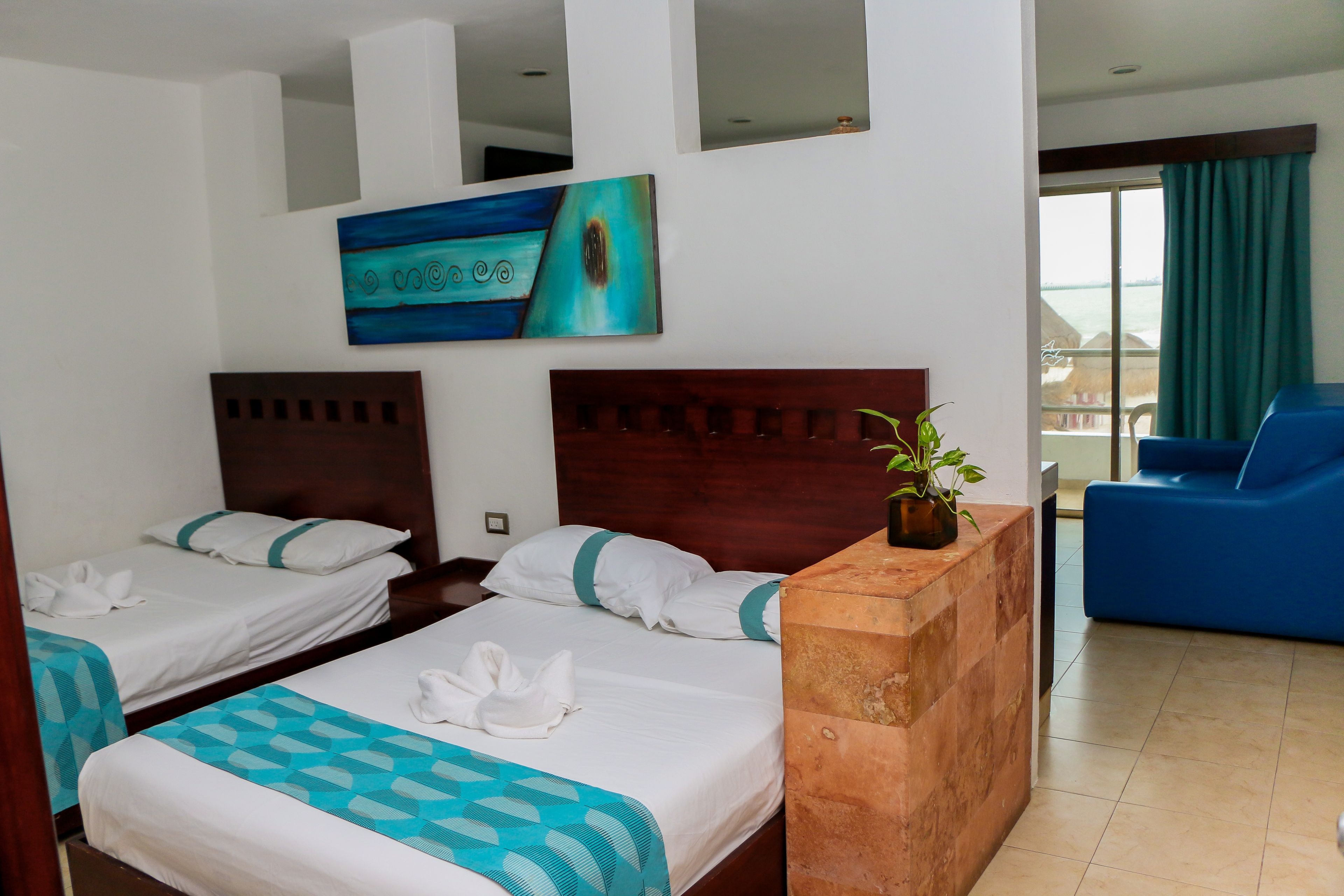 A hotel room with two beds, a wooden headboard, a painting, a potted plant, and a blue chair.
