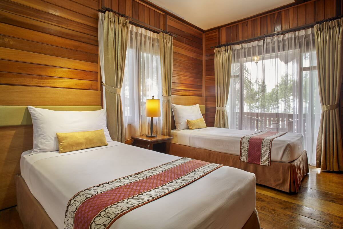 A hotel room with two beds, wooden paneling on the walls, and large windows with curtains.
