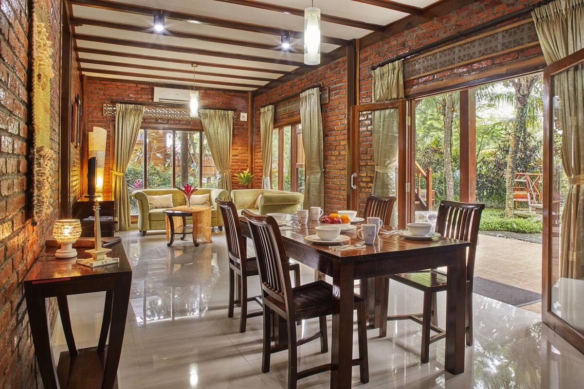 A dining area with a brick wall, wooden ceiling, and large windows. There are dining tables, chairs, and a lamp.