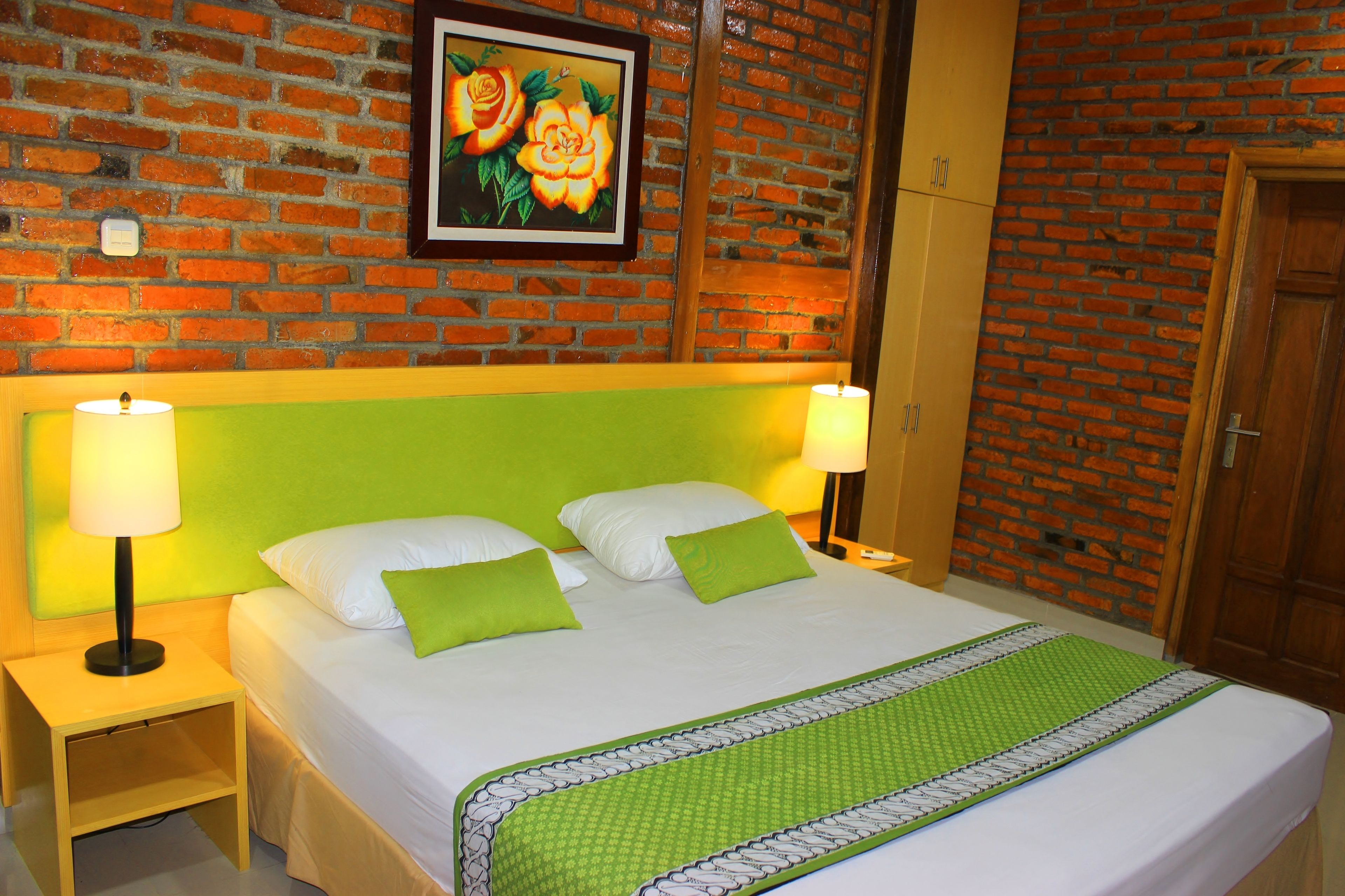A hotel room with a brick wall, a green headboard, a bed with white and green bedding, and a framed picture above the bed.