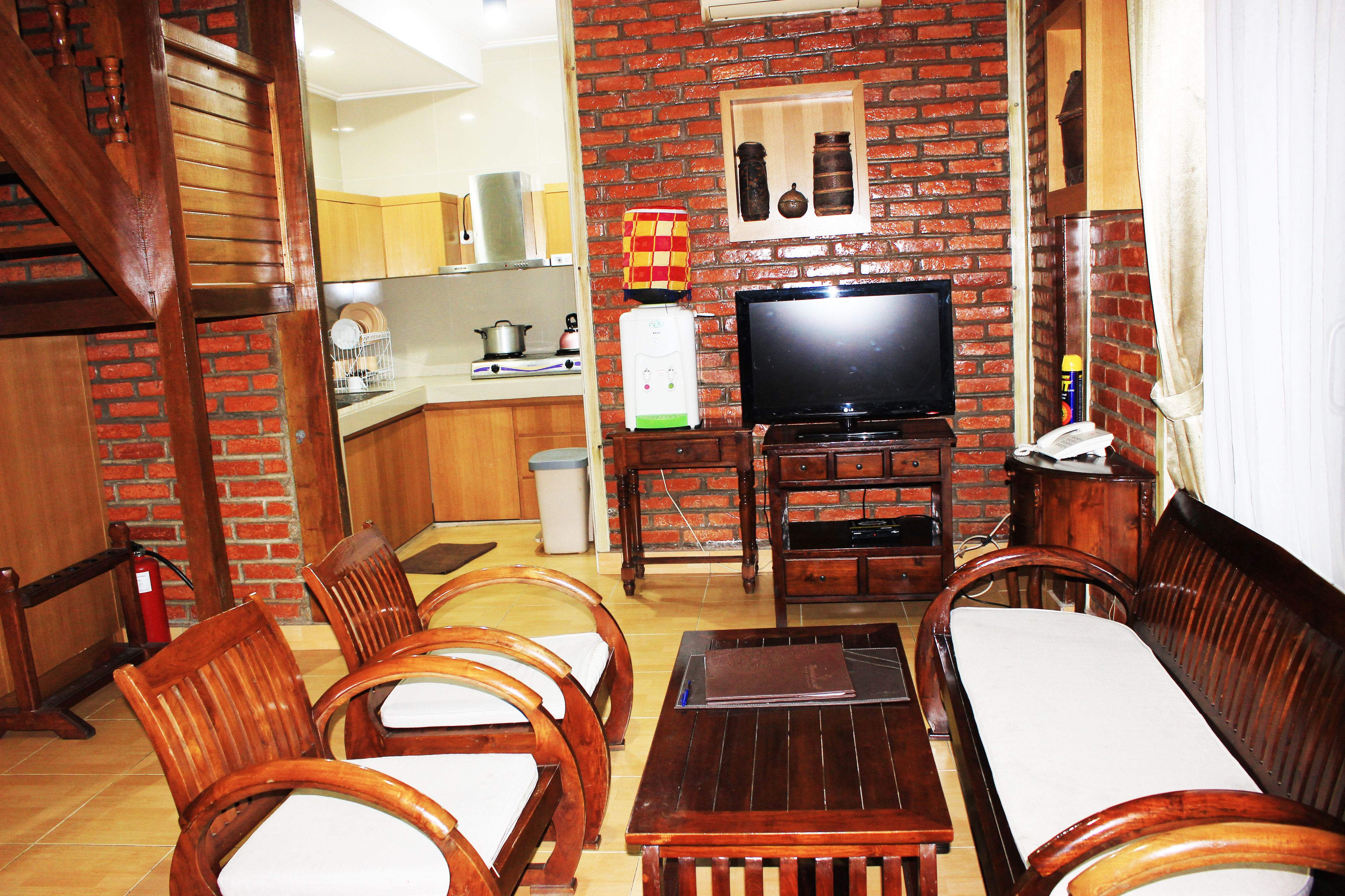 A living room with a brick wall, wooden furniture, a television, and a kitchen in the background.