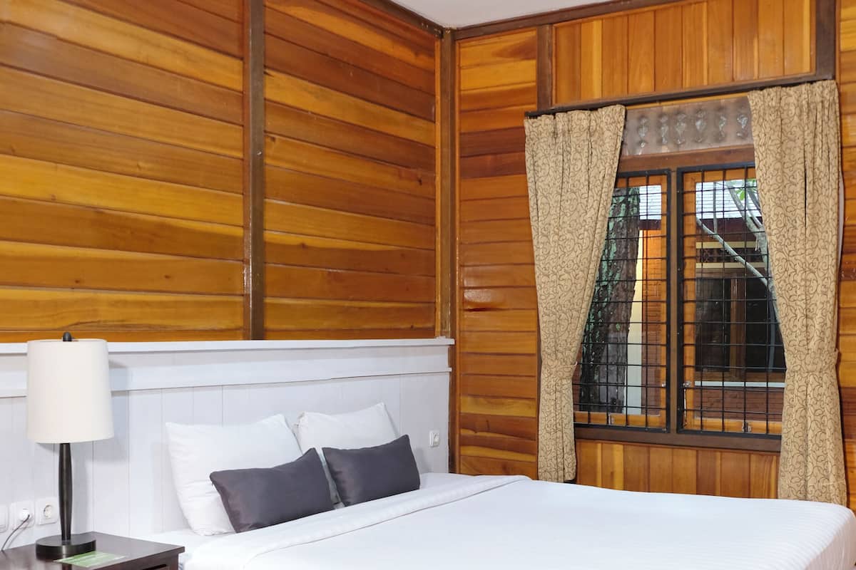 Ein Schlafzimmer mit einer holzverkleideten Wand, einem Bett mit weißer Bettwäsche, einem Nachttisch mit Lampe und einem Fenster mit gemusterten Vorhängen.