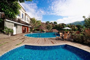 A clear blue swimming pool with wooden deck and lounge chairs, surrounded by lush greenery and a view of a hillside.