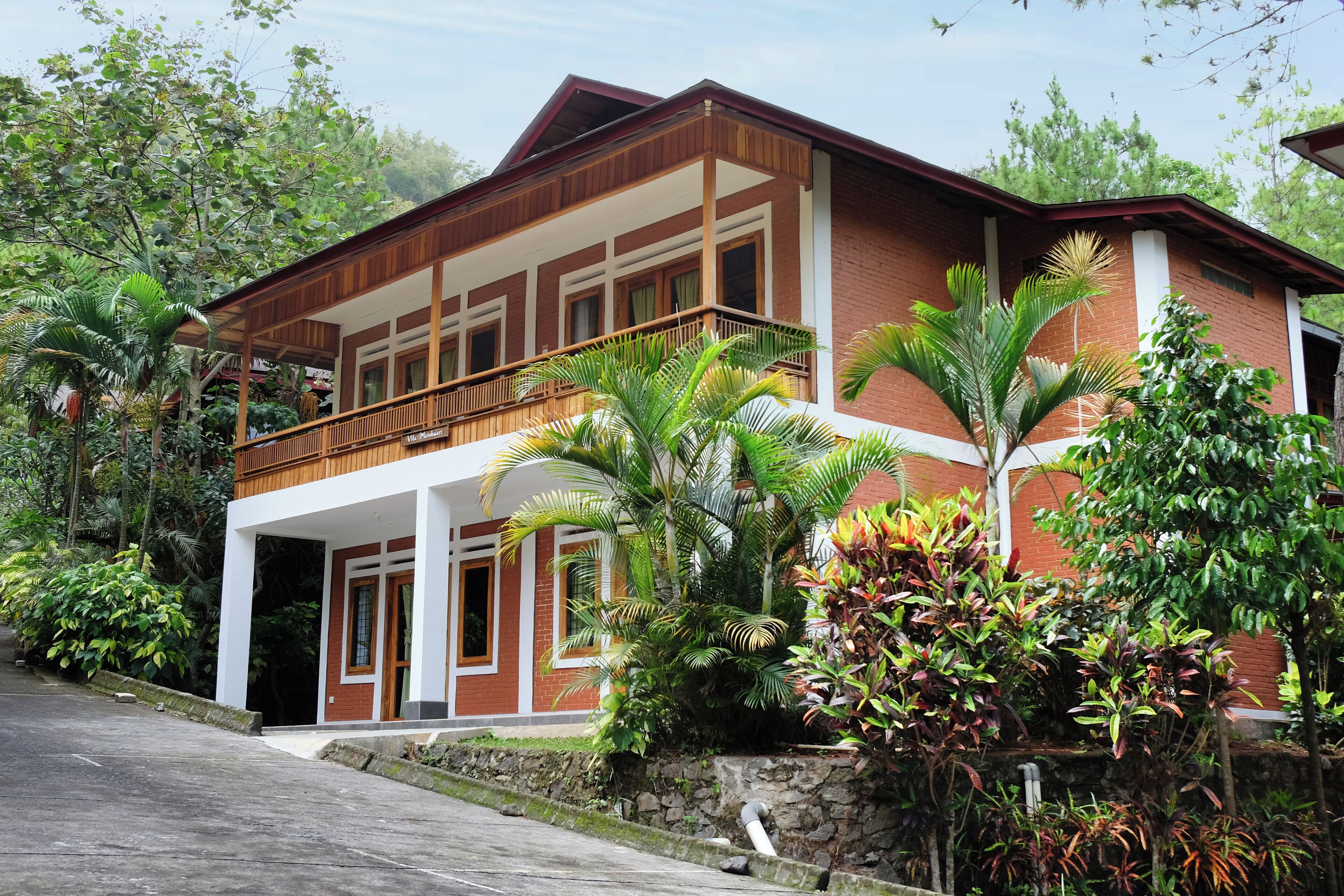 A two-story house with a balcony, surrounded by lush greenery and a stone wall.