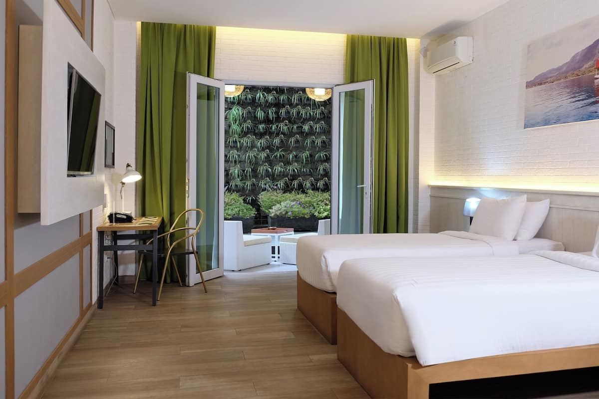 Ein Hotelzimmer mit zwei Betten, einem Schreibtisch mit Stuhl, einem kleinen Tisch und Blick auf Grün durch eine offene Tür.