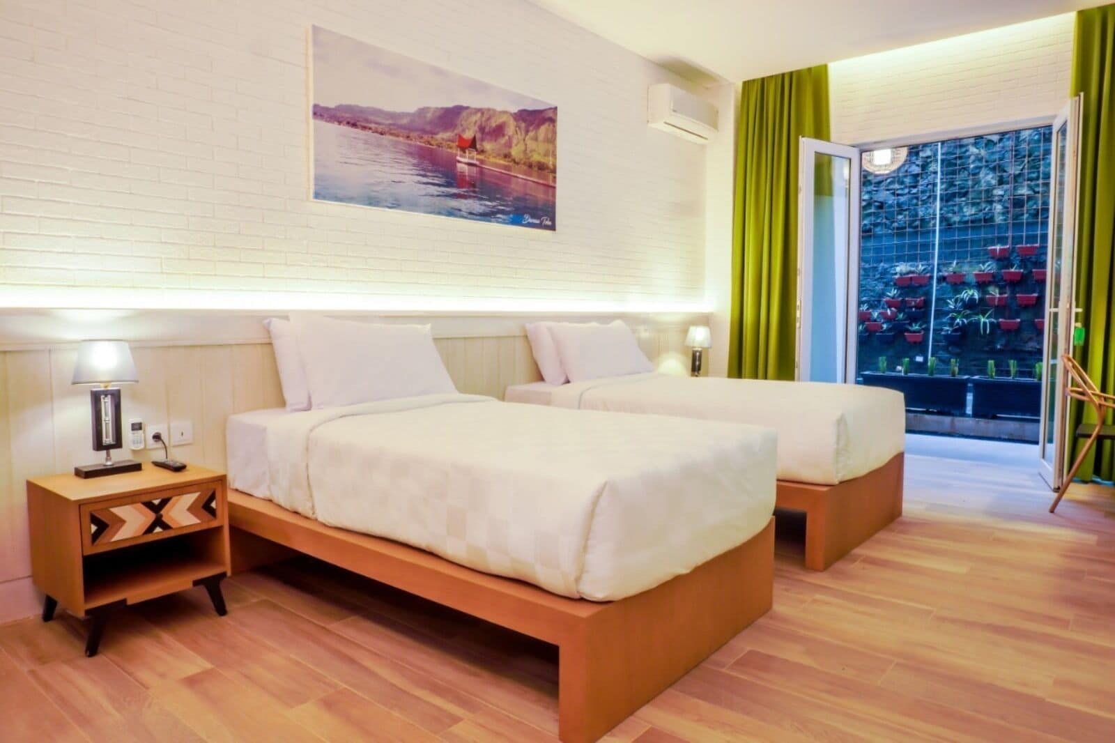 A hotel room with two beds, a nightstand with a lamp, a painting on the wall, and a view of a balcony with plants.