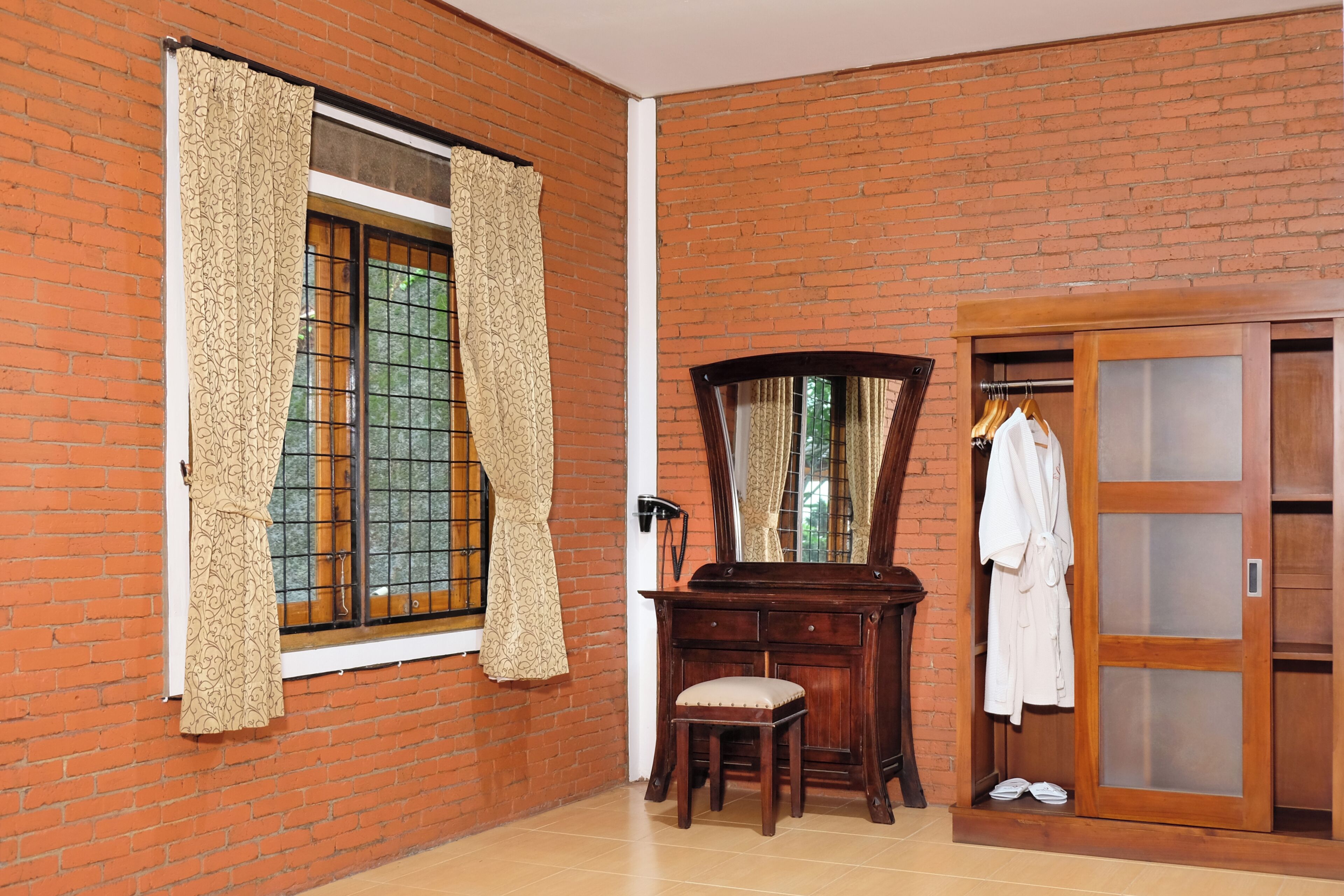 A room with a brick wall, a wooden desk with a chair, a mirror, and a wardrobe with a robe and slippers.
