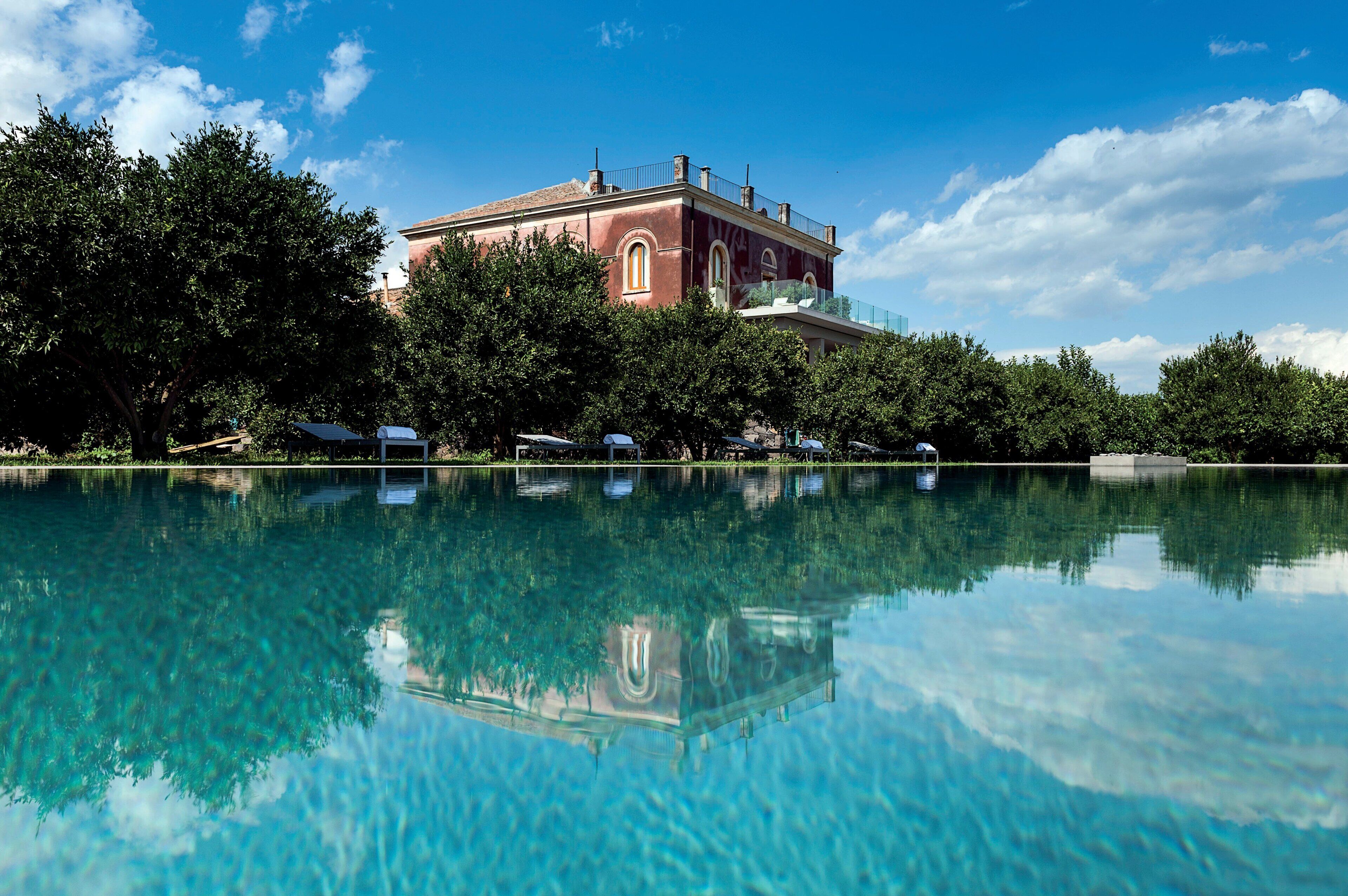 A clear pool reflecting a building with arched windows and a red facade, surrounded by lush greenery.