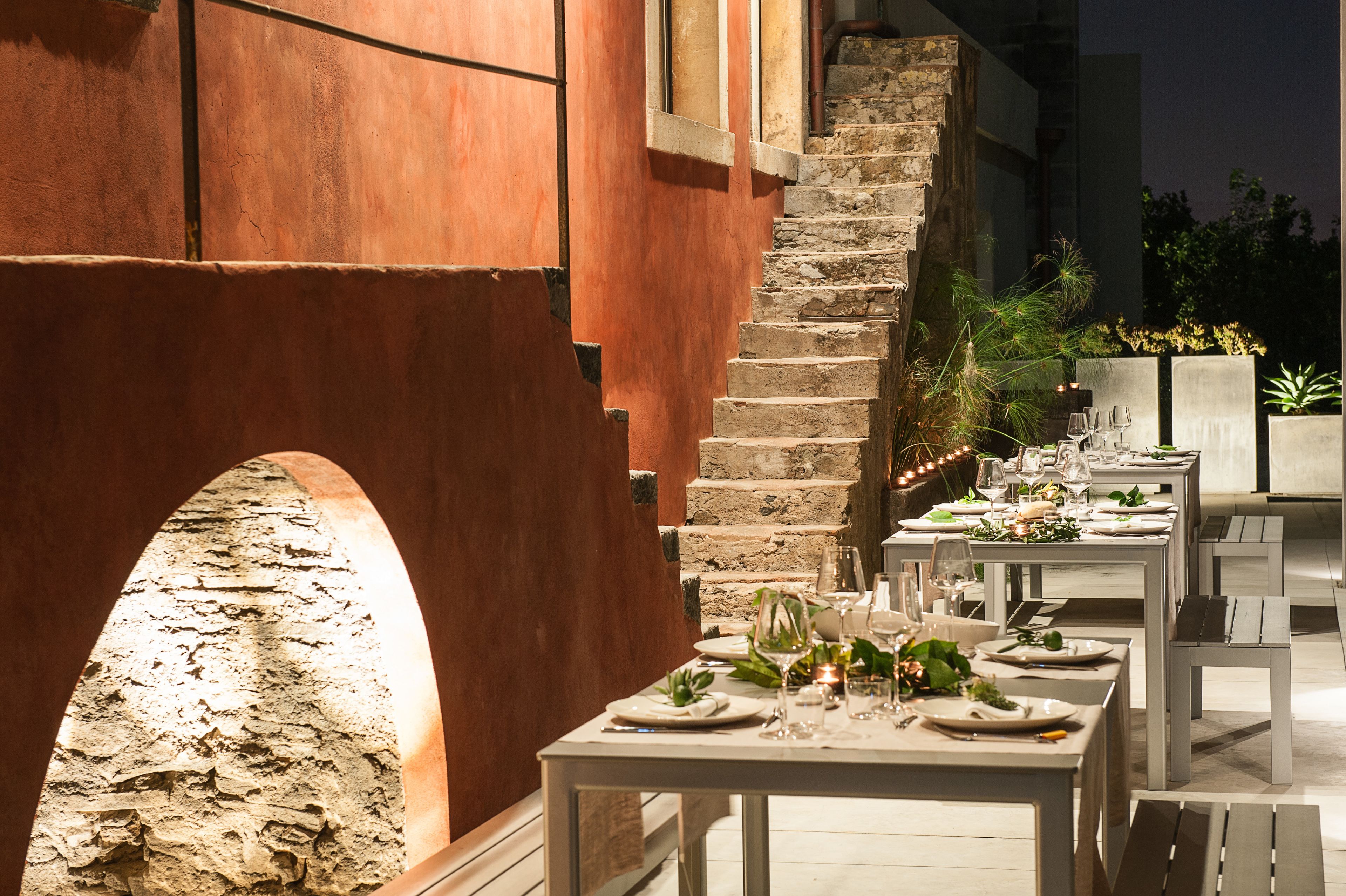 Un espace de restauration en plein air doté de murs en pierre et d’un escalier, avec des tables dressées pour le repas, décorées de bougies et de plantes.