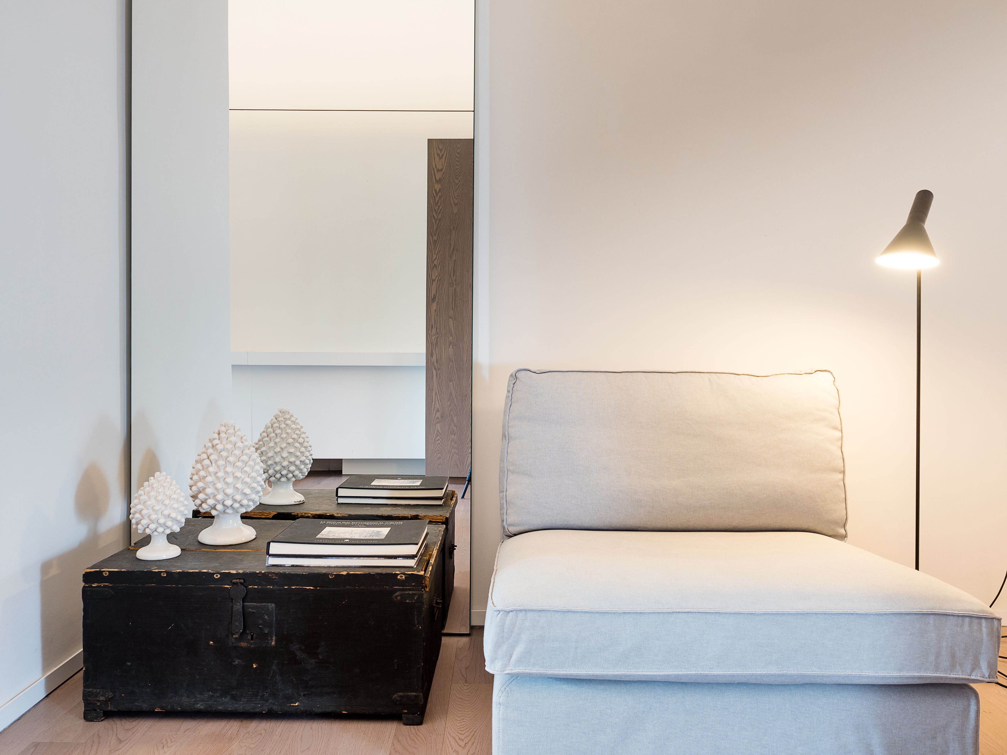 Een minimalistisch ingerichte kamer met een lichtgrijze bank, een zwarte koffer met decoratieve spullen, en een vloerlamp.