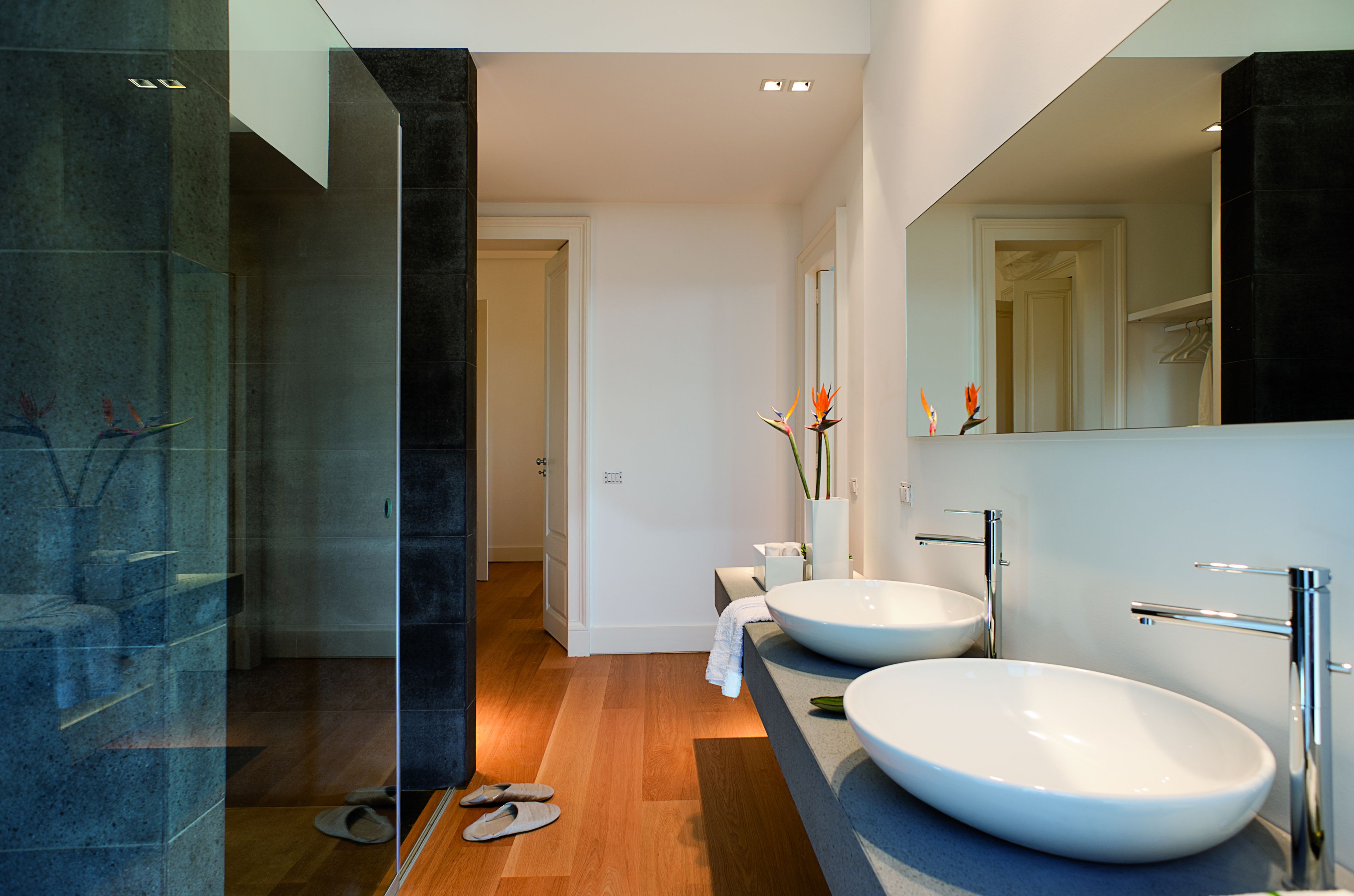 Un baño moderno con dos lavamanos grandes blancos, una cabina de ducha de vidrio y suelo de madera.