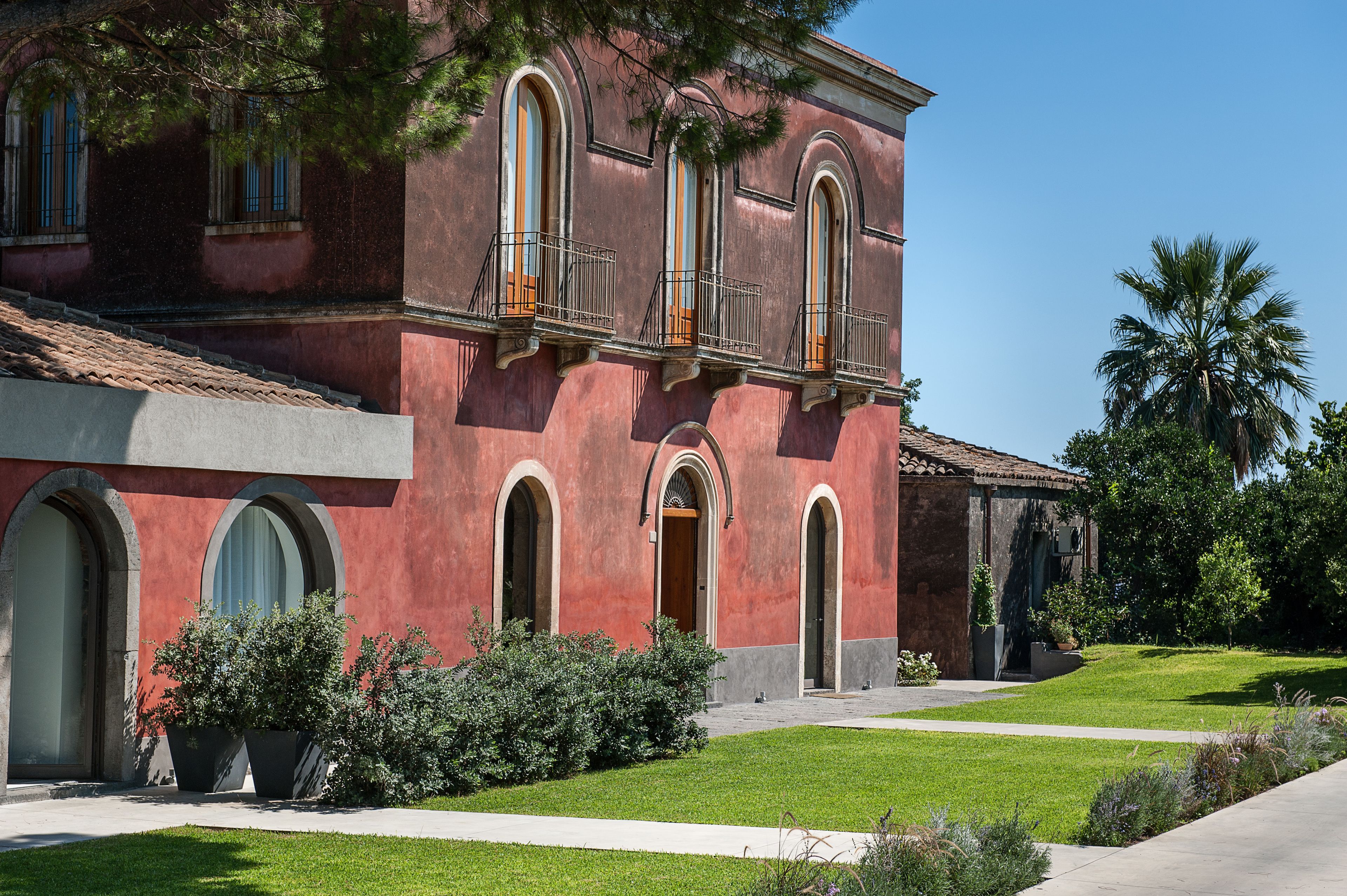 A red building with arched windows and balconies, surrounded by greenery and a clear sky.
