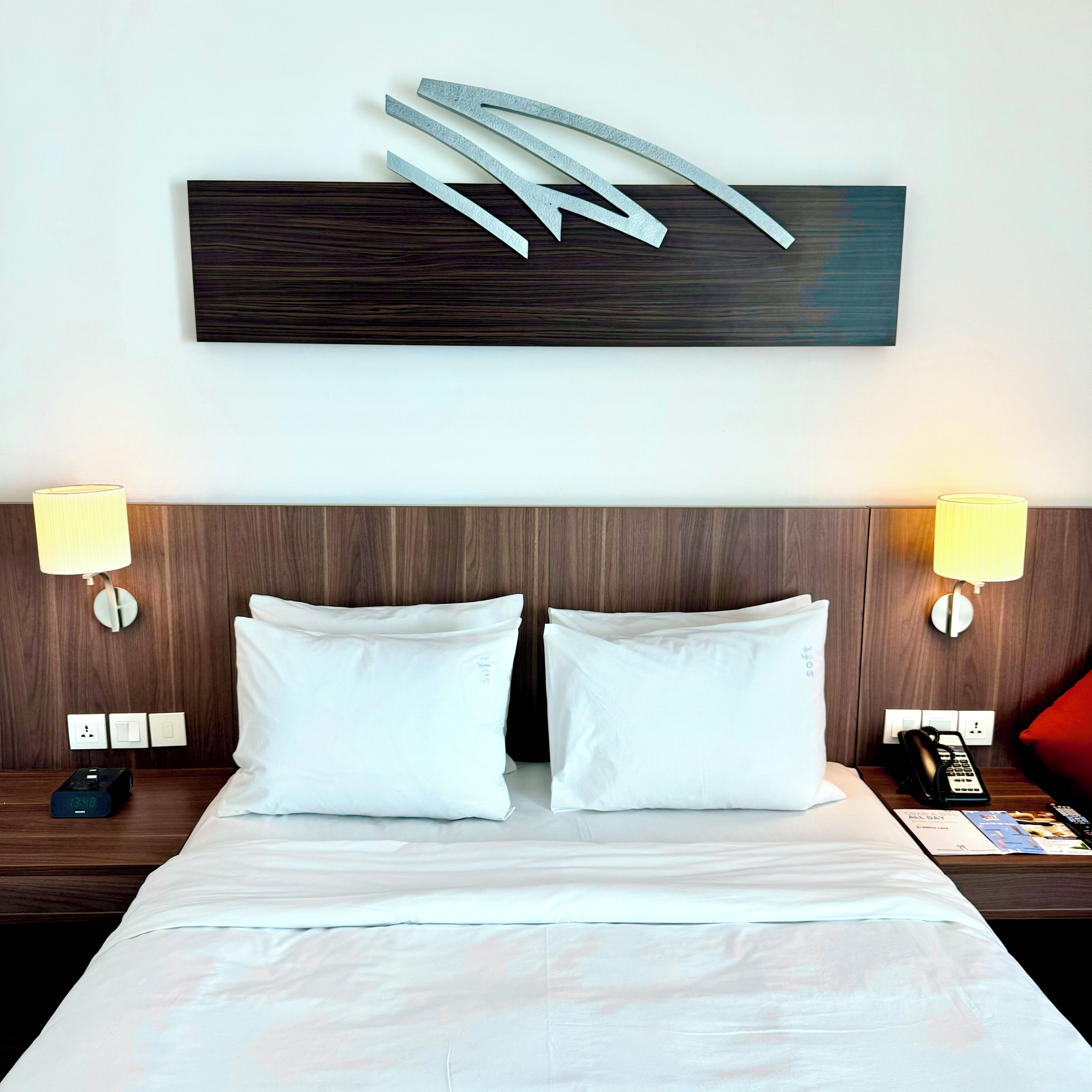 Habitación de hotel con cabecera de madera, dos almohadas blancas, dos lámparas de noche, teléfono junto a la cama y una lámpara de pared.