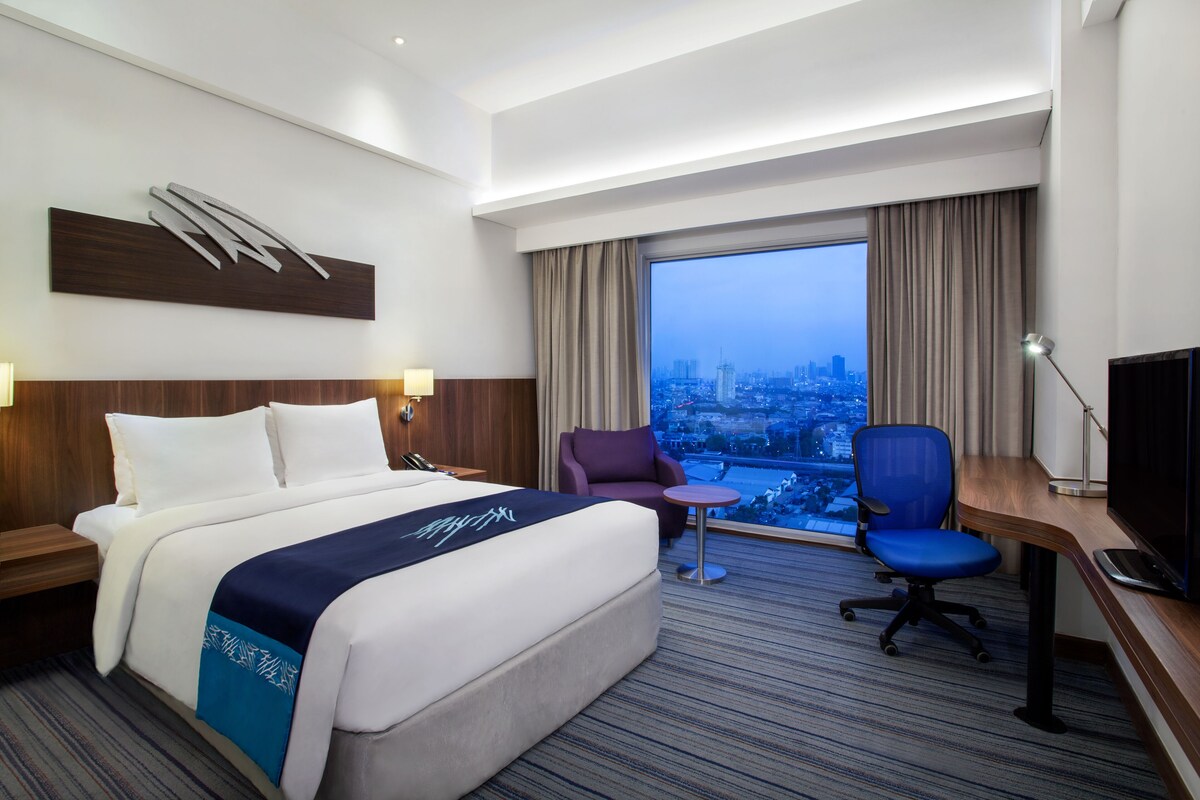 A hotel room with a large bed, a desk with a chair, a TV, and a view of a cityscape through a large window.