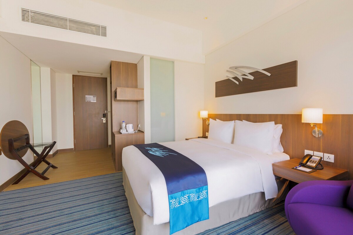 A hotel room with a bed, bedside table, chair, and a desk with a lamp.