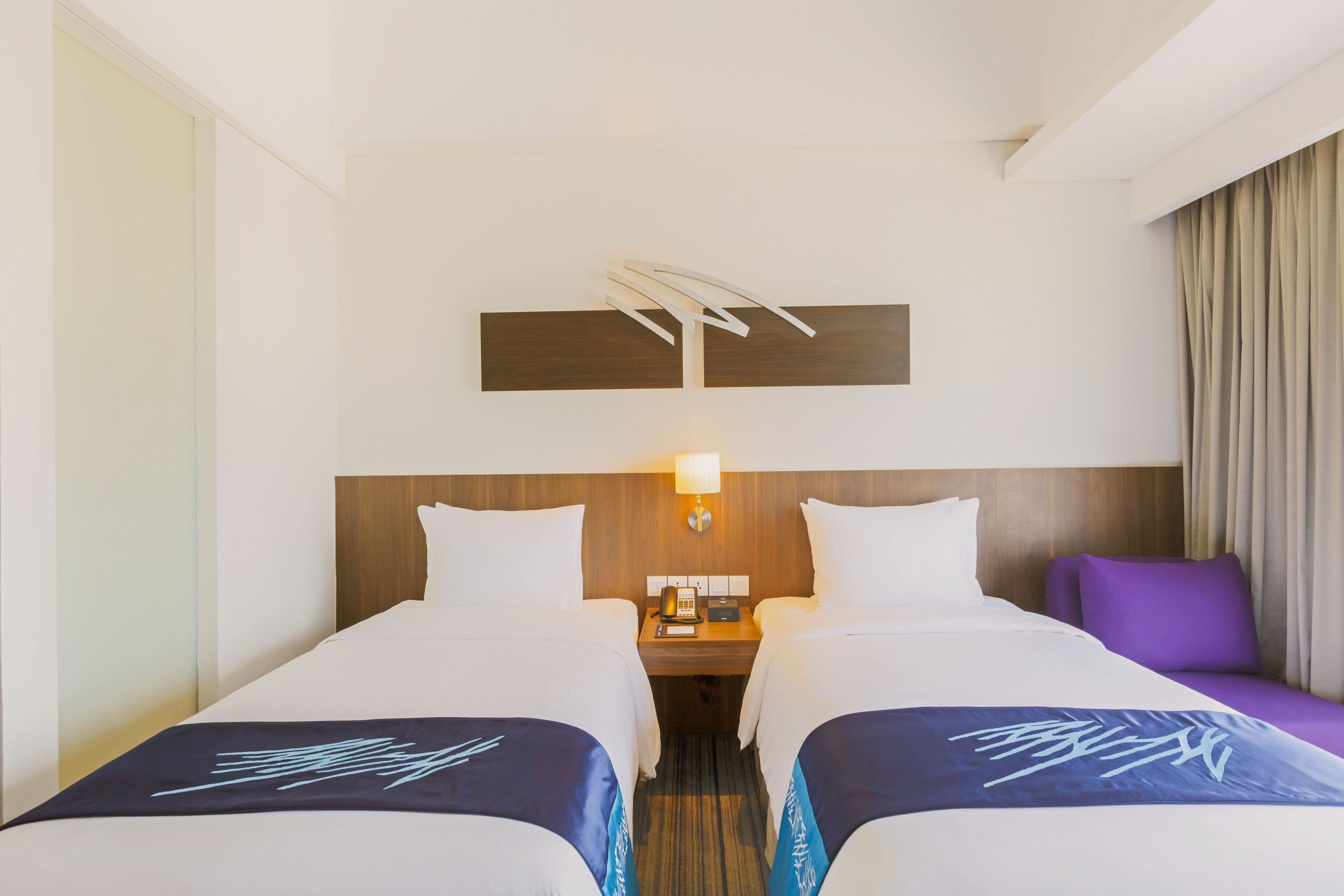 Quarto de hotel com duas camas, uma mesinha de cabeceira, uma luminária e uma luminária fixa na parede.
