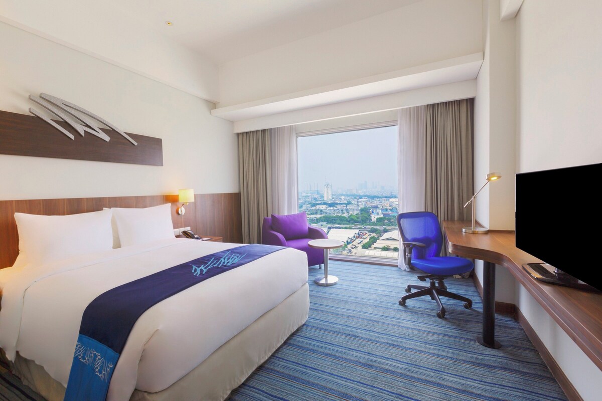 A hotel room with a large bed, a desk with a TV, a chair, and a view of a cityscape.