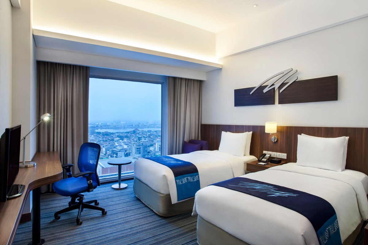 A hotel room with two beds, a desk, a chair, a TV, and a large window with a city view.