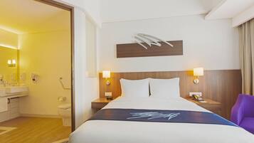 A hotel room with a bed, two bedside lamps, a wooden headboard, a bathroom with a sink and toilet, and a purple chair.